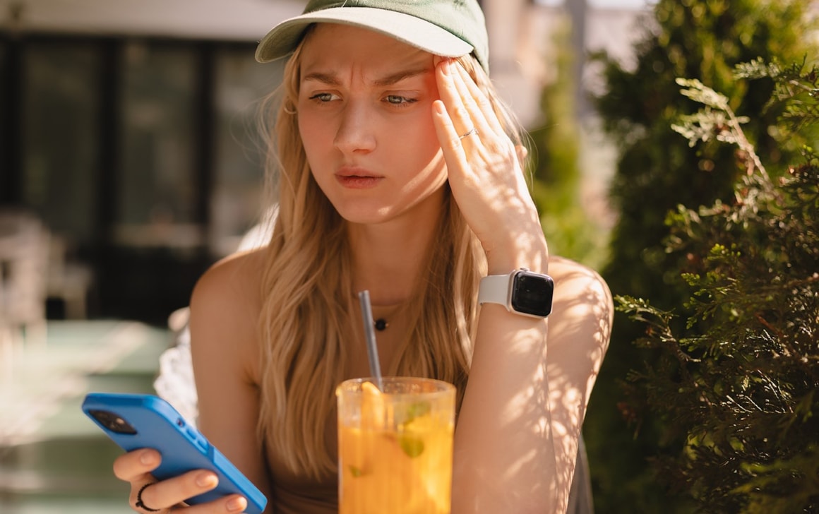 En jente som sitter ute i solen og erger seg over det hun ser på mobilen.